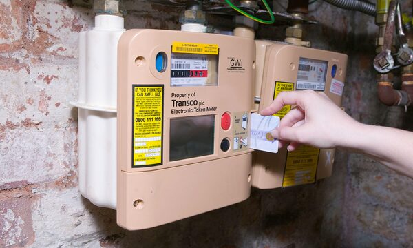A hand inserting a payment card into a Transco electronic token gas meter mounted on a brick wall