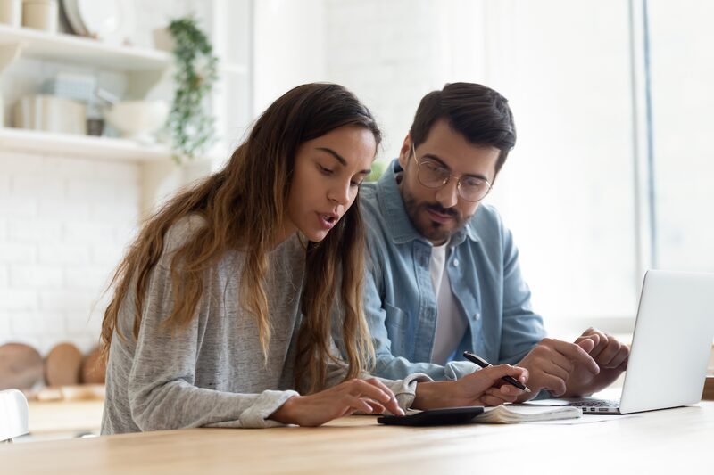 A couple at a kitchen table using a calculator, notebook and laptop to work out household bills