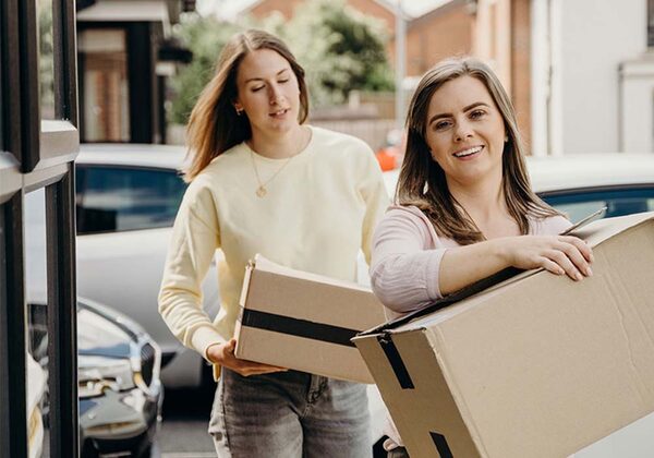 Two women carrying cardboard moving boxes outside a house