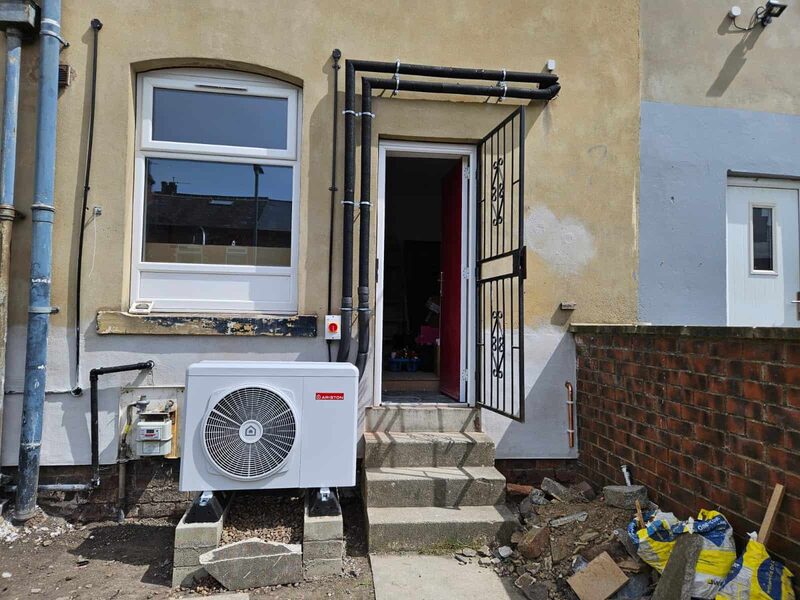 A white Ariston air source heat pump installed on blocks outside the back door of a rendered terraced house, with pipework running up the wall and rubble on the ground