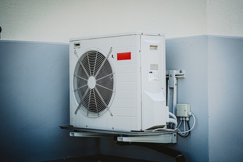 An air source heat pump outdoor unit mounted on a wall bracket against a painted exterior wall