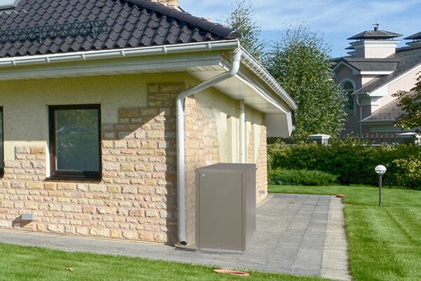 A grey outdoor heating oil tank installed beside the brick wall of a house on a paved path