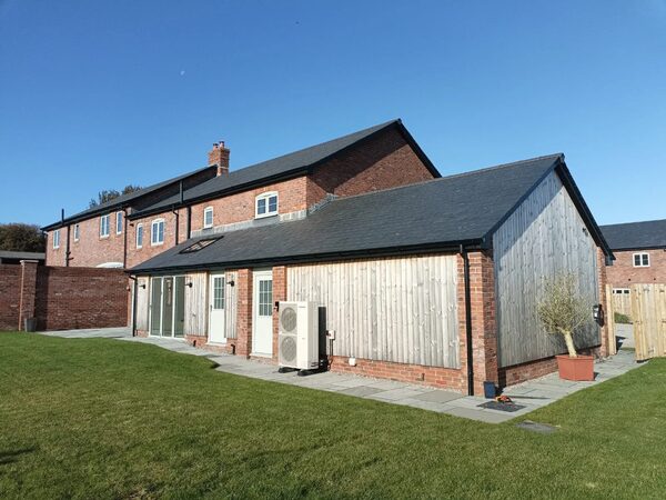An air source heat pump unit installed on the exterior wall of a modern brick and timber-clad house with a lawn in front
