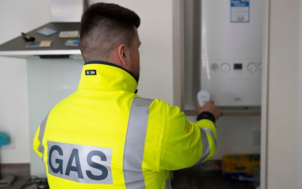 A gas engineer in a high-visibility GAS jacket servicing a wall-mounted boiler in a home kitchen