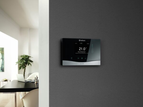 A Vaillant sensoHOME smart thermostat mounted on a grey wall showing 21.0°C, with a dining area in the background