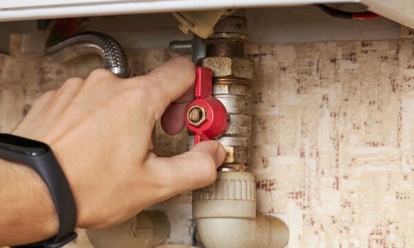 A hand turning a red-handled valve on brass plumbing pipework under a boiler
