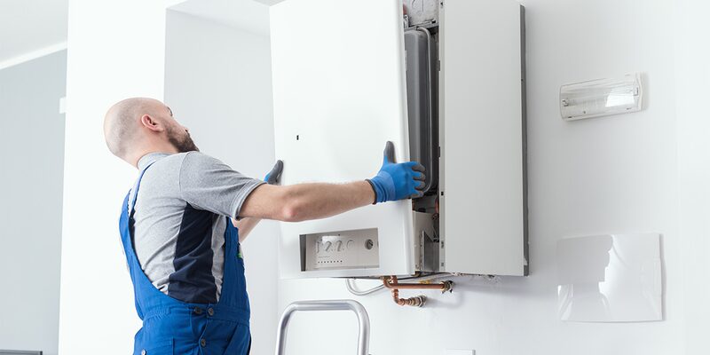 A boiler engineer in blue overalls and gloves removing the casing from a wall-mounted combi boiler