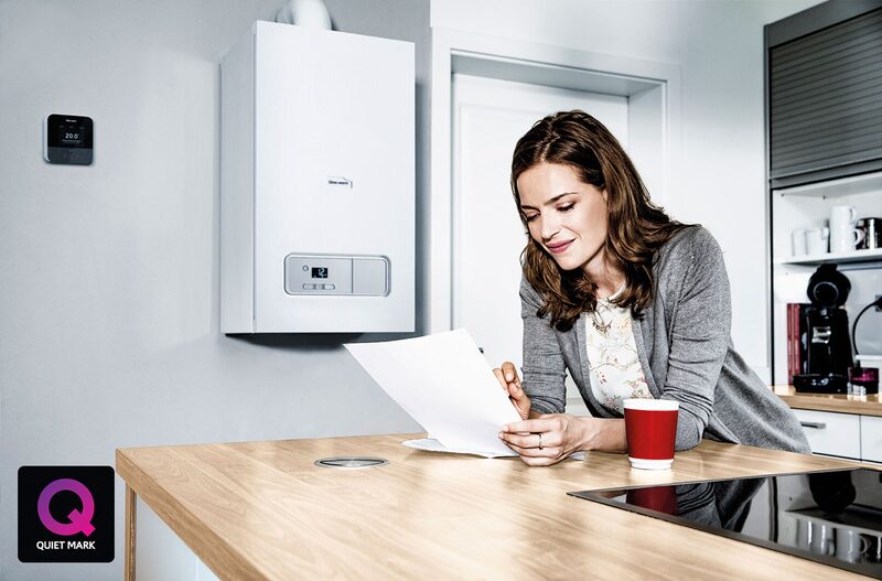 A woman reading a document at a kitchen counter next to a wall-mounted Glow-worm combi boiler with a thermostat, with a Quiet Mark logo in the corner