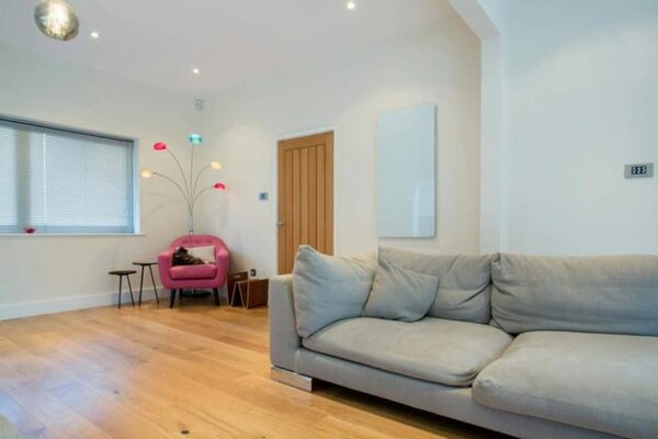 A white infrared heating panel mounted high on the wall of a modern living room with a grey sofa and pink armchair