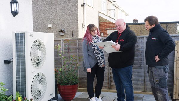 An installer in a Grant UK jacket shows paperwork to a homeowner beside an air source heat pump unit outside a house