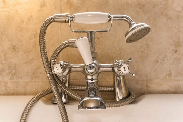 A chrome bath shower mixer tap with a handheld shower head and hoses on a white bathtub against a beige tiled wall