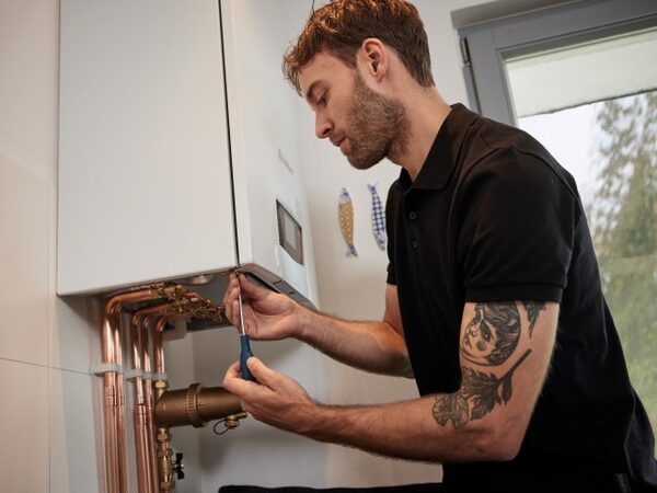 A heating engineer using a screwdriver to work on a wall-mounted white boiler with copper pipework in a home
