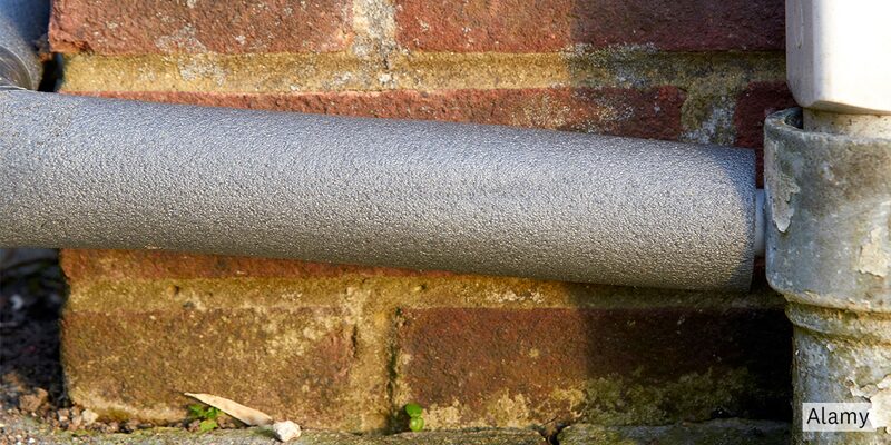 A frosted-over condensate pipe running along an exterior brick wall outside a house