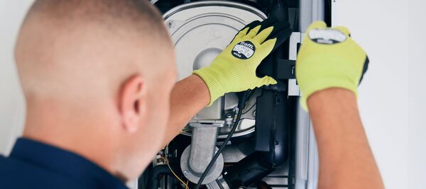 An engineer in yellow gloves servicing the internal components of a wall-mounted gas boiler