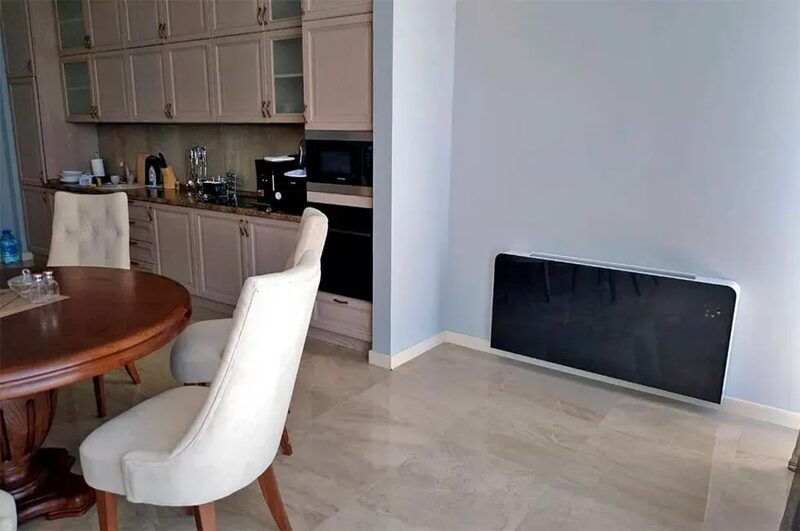 A wall-mounted black panel heater installed low on the wall of an apartment dining area with a wooden table, cream chairs and kitchen units