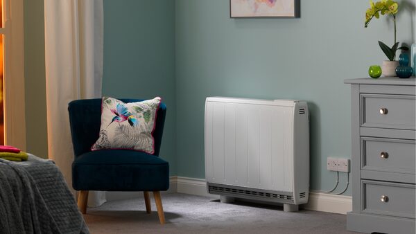 A white electric storage heater installed on the wall of a furnished bedroom with an armchair and chest of drawers