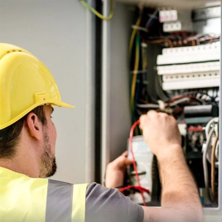 An electrician in a yellow hard hat and hi-vis vest testing wiring in a consumer unit with a multimeter