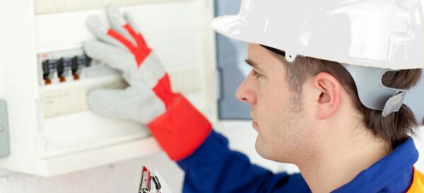 A technician in a white hard hat and gloves testing a wall-mounted electrical distribution board