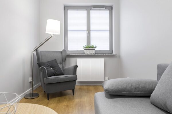 A white electric radiator installed on the wall under a window in a grey living room with an armchair and sofa