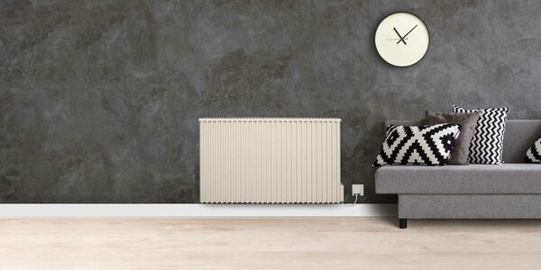 A cream Fischer Classic electric radiator mounted on a dark grey wall beside a grey sofa with patterned cushions and a wall clock