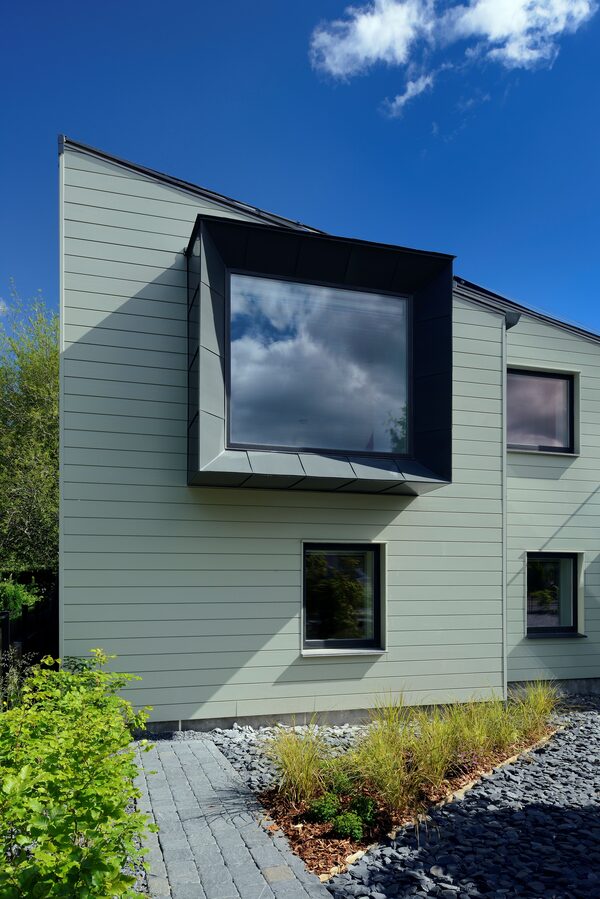 A modern energy-efficient house in Scotland with pale green timber cladding, large dark-framed windows and a gravel garden under a blue sky