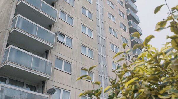 A tall concrete block of flats with balconies and windows, partly obscured by a leafy shrub in the foreground