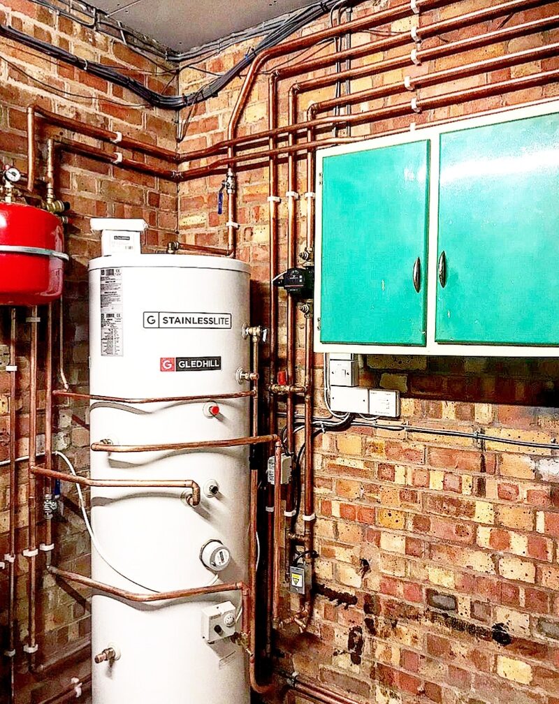 A white Gledhill StainlessLite hot water cylinder installed against a brick wall with copper pipework and a red expansion vessel