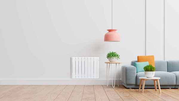 A white Haverland electric radiator mounted on a plain wall in a bright living room with a grey sofa, side tables and a pink pendant lamp
