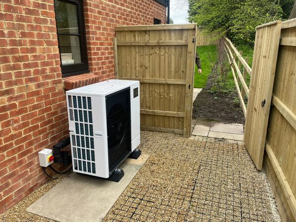 An air source heat pump unit installed outside a brick house beside a wooden garden gate