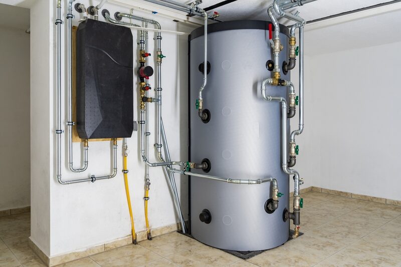 A large grey cylindrical hot water thermal storage cylinder with extensive copper pipework, valves and a wall-mounted unit in a plant room