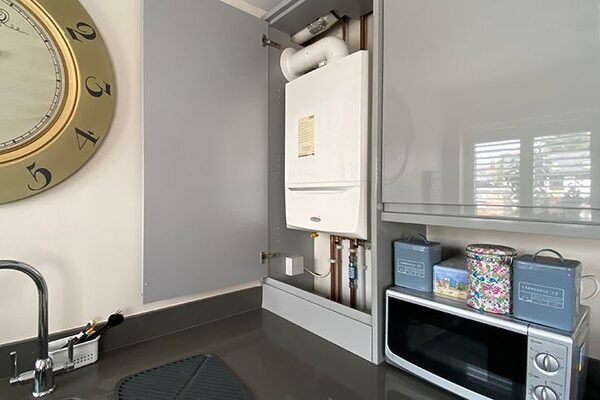 A white wall-mounted gas combi boiler installed inside a grey kitchen cupboard with the door open