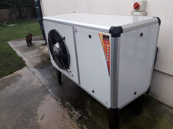 An air source heat pump unit installed outside a house on a concrete path, with a cat walking away in the background
