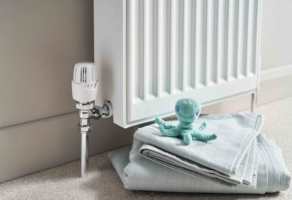 A white thermostatic radiator valve (TRV) fitted to the pipework beneath a white panel radiator, with folded towels and a toy octopus on the carpet beside it