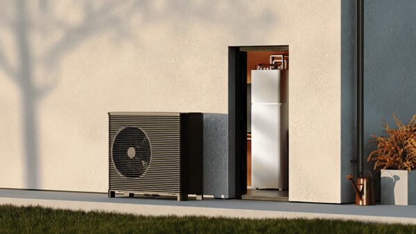 A dark heat pump unit installed outside a house wall next to an open utility door with a hot water cylinder inside