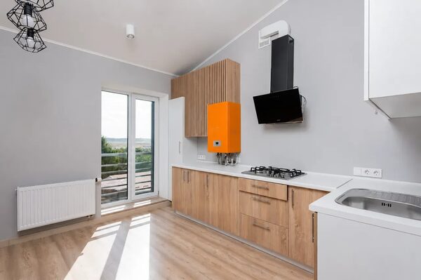 An orange wall-mounted gas boiler installed in a modern kitchen with wooden cabinets, a gas hob, cooker hood and a white radiator