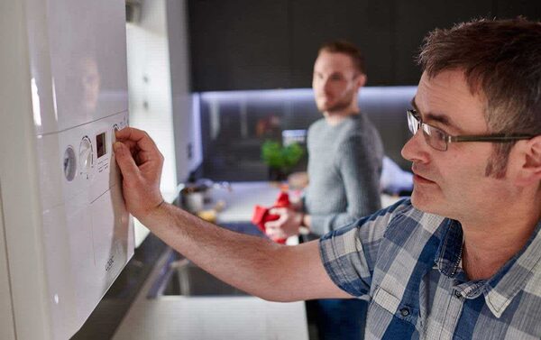 A man adjusting the controls of a wall-mounted Ideal combi boiler in a kitchen while another man stands behind him