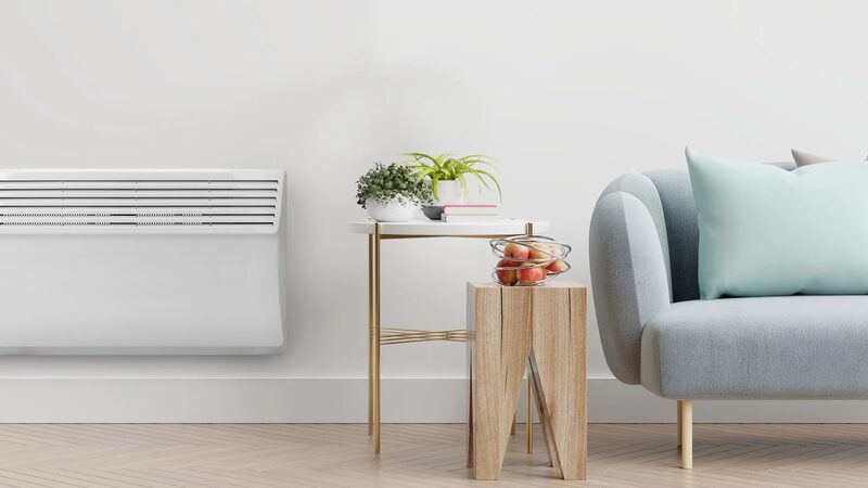A white wall-mounted panel heater in a bright living room beside a grey sofa and side table