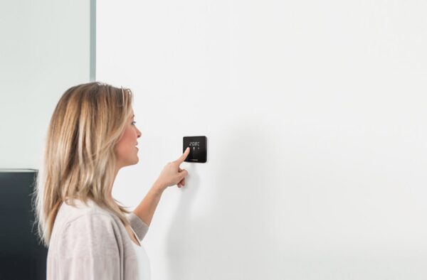 A woman adjusting a black wall-mounted smart thermostat on a white wall