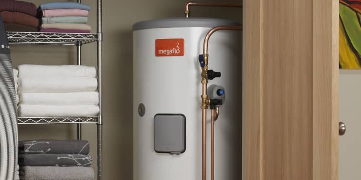 A white Megaflo unvented hot water cylinder with copper pipework installed in an airing cupboard next to shelving with folded towels
