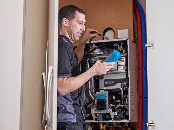 A technician servicing a wall-mounted Navien gas boiler in an airing cupboard, holding a blue flue gas analyser