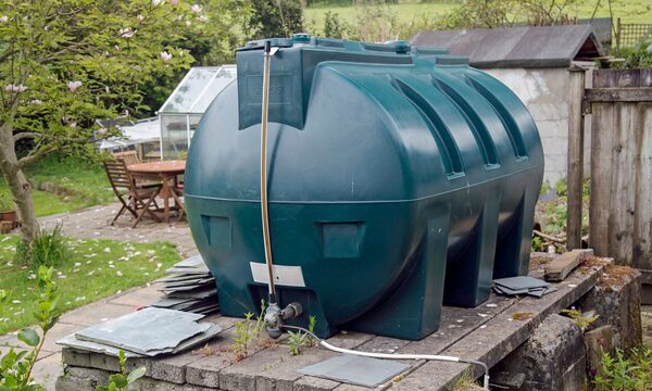 A large green plastic heating oil tank installed on a stone platform in a garden with patio furniture and a shed behind.