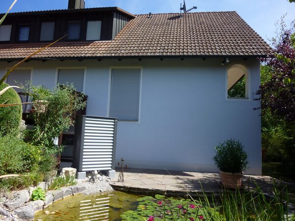 An outdoor heat pump unit installed beside the exterior wall of a house with a garden pond in the foreground