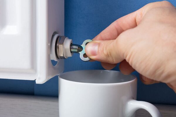 A hand using a radiator bleed key on a white radiator's bleed valve with a white mug below to catch drips