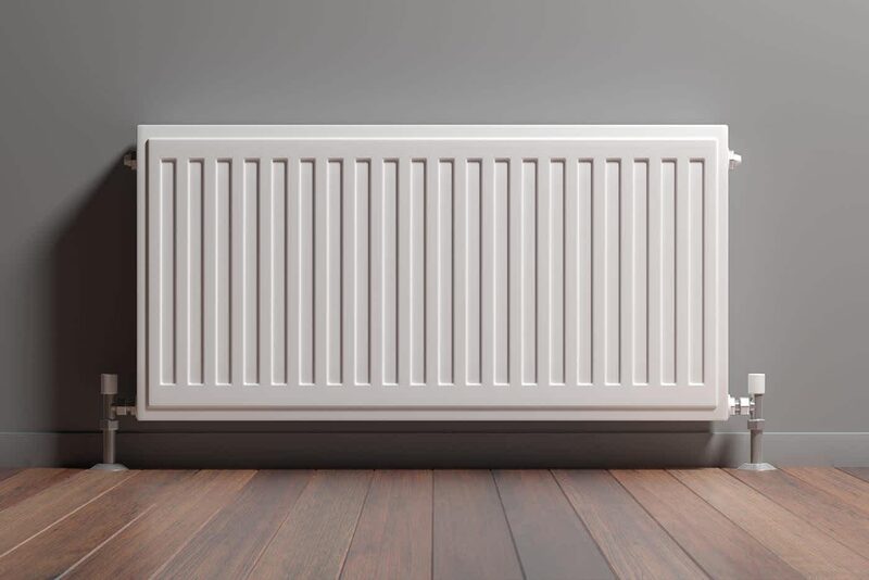 A white panel radiator mounted on a grey wall above a wooden floor, with visible pipework and valves at each side