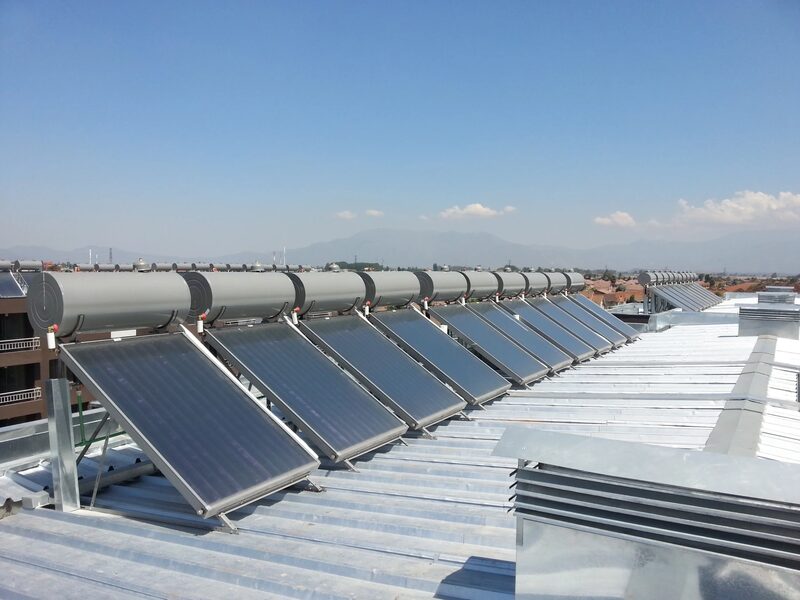 A row of solar thermal collectors with cylindrical hot water tanks installed on a large flat metal roof