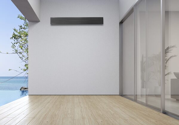 A slim black Sundirect SC-Plus infrared panel heater mounted high on a white wall of a modern room with wooden flooring and glass doors opening to a sea view