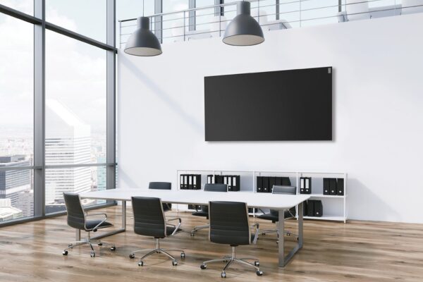 A black flat panel wall heater mounted high on the wall of a modern office meeting room with a white table, chairs and pendant lights