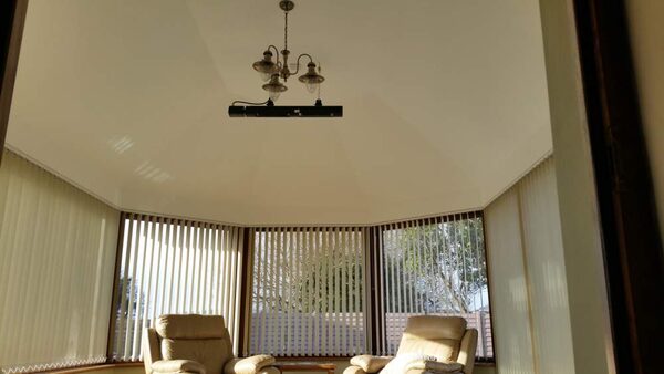 A black electric radiant heater mounted on the ceiling of a conservatory with armchairs and vertical blinds