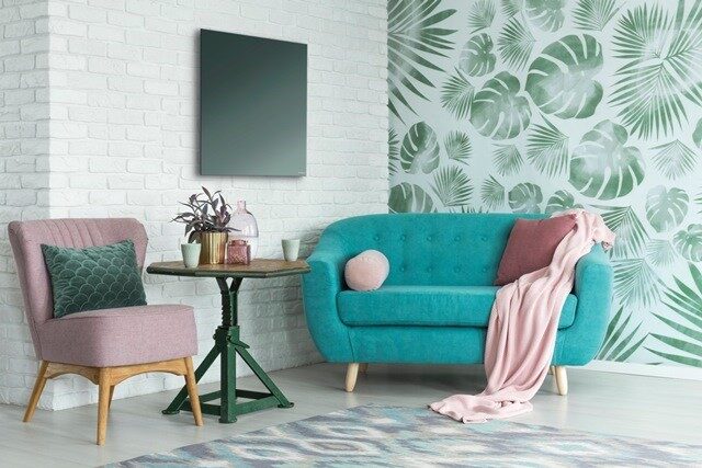 A dark square infrared heating panel mounted on a white brick wall above a pink armchair and teal sofa in a styled living room