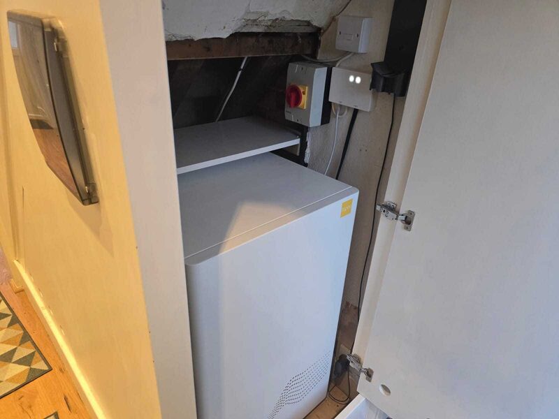 A white tepeo heat battery installed in a cramped cupboard under stairs with electrical isolator switch above